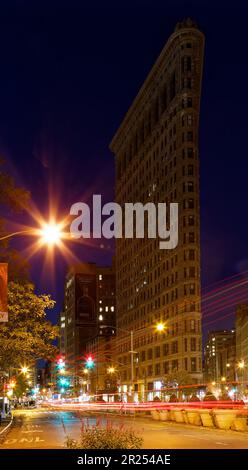 Il Flatiron Building si illumina caldo sotto i semafori sotto un cielo indaco prima dell'alba; il traffico sparso lascia sentieri leggeri nella foto di esposizione al tempo. Foto Stock