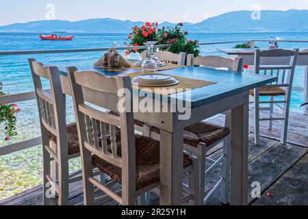 Una vista accattivante di un Ristorante Taverna vuoto presso il pittoresco lungomare, caratterizzato da invitanti sedie e tavoli in legno Foto Stock