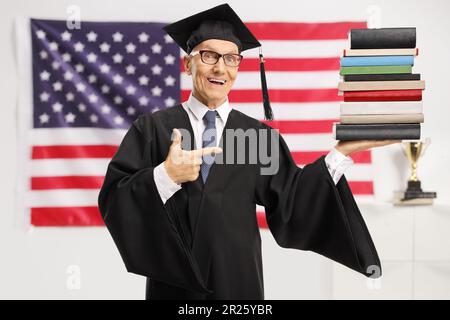 Uomo anziano che indossa un abito da laurea e che tiene un mucchio di libri davanti alla bandiera degli Stati Uniti Foto Stock