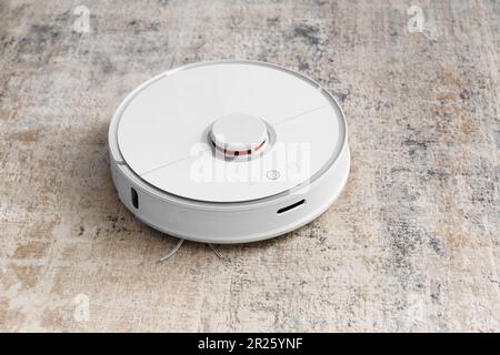 Aspirapolvere robot bianco sul tappeto nel soggiorno vicino al divano. Pulire l'appartamento con un moderno aspirapolvere intelligente. Foto Stock