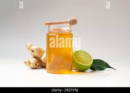 Miele biologico in vaso trasparente di vetro, zenzero, lime, noci e un cucchiaia di legno su fondo bianco. Cucchiaio per miele e miele fresco Foto Stock