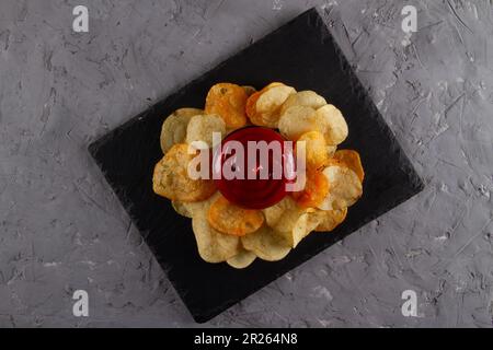 Vista dall'alto di un mucchio di patatine fritte con salsa di immersione rossa sul piano di servizio in ardesia nera sul tavolo in gesso grigio. Elegante e divertente piatto Foto Stock