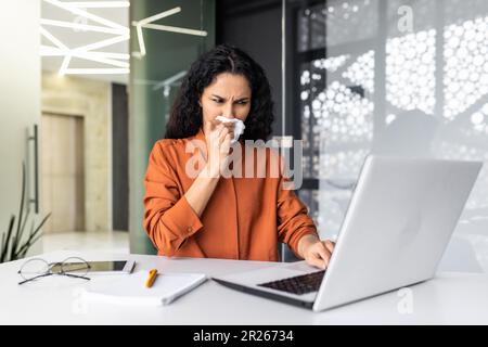 Una giovane donna musulmana lavora in ufficio su un computer portatile, si siede a un tavolo e si sente male. Ha un naso che cola, allergie, un virus. Si asciuga il naso con un tovagliolo, starnuti e tosse, ha bisogno di trattamento. Foto Stock