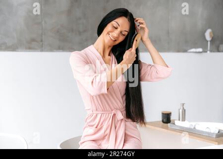 Routine di bellezza femminile. Pettinare i capelli. Bella donna brunetta caucasica, seduta in un elegante bagno moderno, in un accappatoio, pettina i capelli lunghi con un pettine di legno, sorride Foto Stock