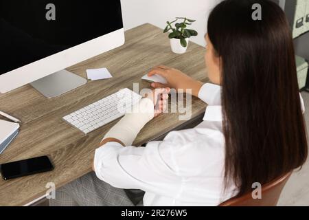 Giovane donna con braccio avvolto in bendaggio medico seduto sul posto di lavoro Foto Stock