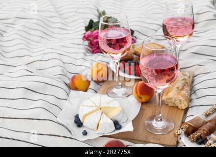 Bicchieri di delizioso vino rosa, fiori e cibo su coperta bianca da picnic Foto Stock