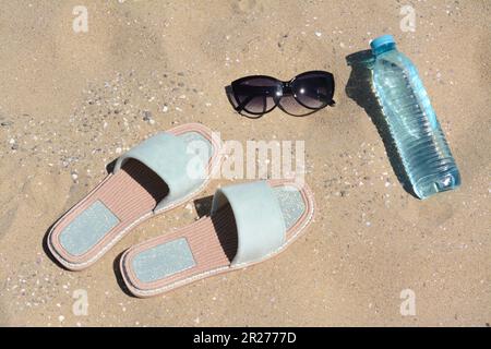 Eleganti occhiali da sole, ciabatte e bottiglia d'acqua sulla sabbia. Accessori da spiaggia Foto Stock