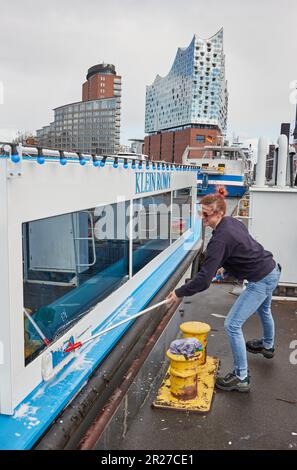Amburgo, Germania. 16th maggio, 2023. Jaqueline Wegenhaupt, skipper del porto, pulisce la chiatta XXL 'Klein Romy' a Niederhafen. La Sala Filarmonica dell'Elba è visibile sullo sfondo. Credit: Georg Wendt/dpa/Alamy Live News Foto Stock