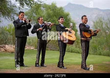 I membri della Mariachi band in abiti tradizionali suonano strumenti musicali (tromba, violino, guitarrón, vihuela) all'esterno in un giardino, Tucson, Arizona Foto Stock