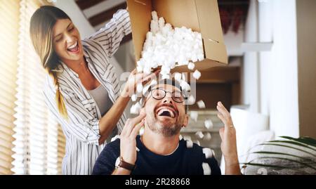 Spostare casa, proprietà e scatola con una coppia sciocca sposata che si divertono mentre giocano nella loro nuova casa insieme. Immobiliare, comico o commedia con un Foto Stock