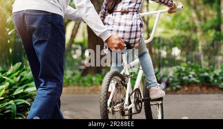 Padre insegna al bambino a fare un giro in bicicletta su un sentiero in un verde parco comunitario all'aperto. Amore, legame e primo piano di un uomo che aiuta la sua bambina su un Foto Stock