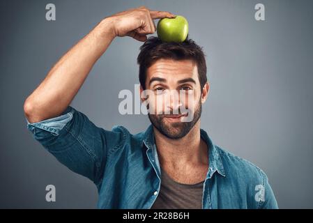 È questo che cosa denominino una dieta equilibrata. Studio shot di un bel giovane uomo che bilancia una mela sulla sua testa su uno sfondo grigio. Foto Stock