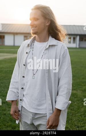 giovane uomo di yoga dai capelli lunghi in camicia di lino e perle sorridente mentre si trova in campagna all'aperto Foto Stock