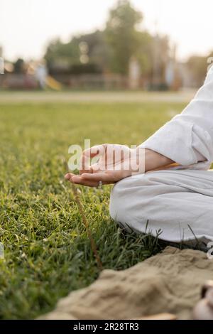 vista ritagliata dell'uomo in vestiti di lino bianco meditando con il gesto di gyan mudra mentre si siede sul campo erboso verde Foto Stock