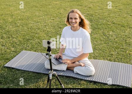 uomo di yoga spensierato che guarda la macchina fotografica mentre seduto con il laptop vicino al treppiede con il telefono cellulare sul prato verde Foto Stock