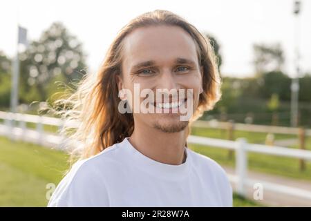 ritratto di felice uomo yoga a capelli lunghi in t-shirt bianca sorridendo alla fotocamera all'aperto Foto Stock