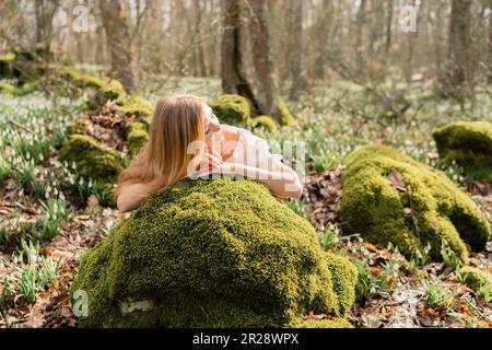 Gocce di neve galanthus biondo. Una ragazza vestita di bianco si stende su una pietra nel muschio in un prato con gocce di neve in una foresta primaverile Foto Stock