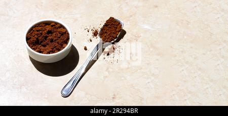 Una ciotola bianca in ceramica e un cucchiaio d'argento riempito di caffè macinato su un tavolo di marmo chiaro in una mattinata di sole. Primo piano di caffè macinato tostato isolat Foto Stock