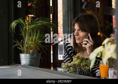 giovane donna transgender latina di etnia argentina, che parla al telefono seduto nel ristorante, guardando fuori dalla finestra e guardando nel di Foto Stock