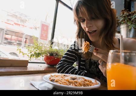 giovane donna transgender latina di etnia argentina in un ristorante seduto con la bocca aperta mangiare spaghetti, concetto di cibo, spazio copia. Foto Stock