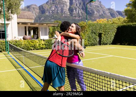 Felice coppia diversa reggendo racchette, abbracciando su rete sul campo da tennis all'aperto soleggiato Foto Stock