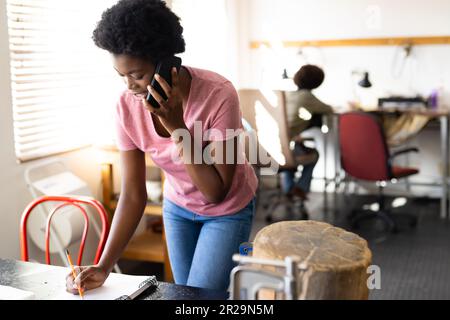 Lavoratrice afro-americana che parla su smartphone e prende appunti al laboratorio di gioielleria Foto Stock