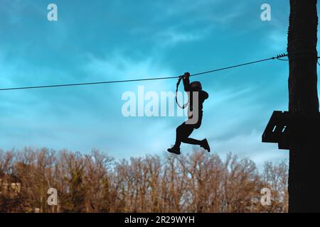 Silhouette di una persona irriconoscibile con zip in un parco avventura. Indossando un casco e un'imbracatura di sicurezza, vengono catturati a metà aria contro un blu Foto Stock