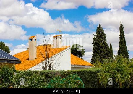 Camini di pietra sui tetti con scandole arancioni contro il cielo blu con nuvole bianche. Una casa di campagna tra cipressi verdi investire in immobili Foto Stock