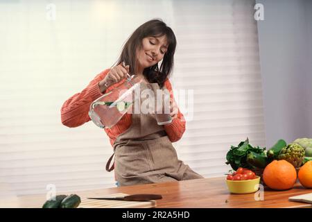 Donna caucasica in un grembiule da cucina siede a un tavolo da cucina, retroilluminata da una grande finestra, e versa l'acqua infusa di cetrioli da una caraffa in un bicchiere. F Foto Stock