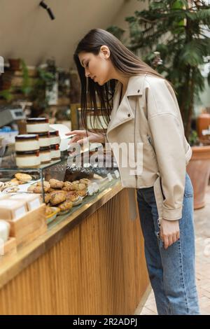 bruna giovane donna con capelli lunghi in giacca di pelle beige e jeans denim in piedi vicino alla torta di esposizione e scegliendo la pasta vicino a vasi di marmellata in moderno Foto Stock