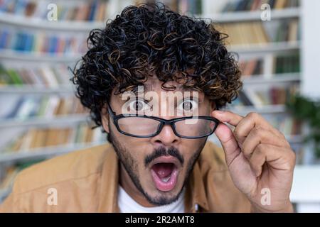 Primo piano. Ritratto di un giovane ispanico in una biblioteca, libreria, che guarda la macchina fotografica di sorpresa, abbassa gli occhiali sul viso. Foto Stock