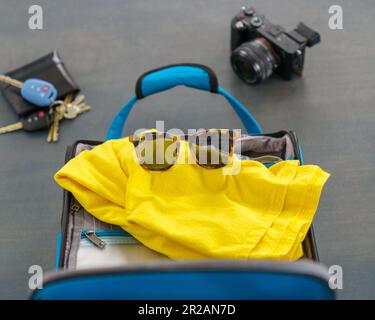 Valigia blu con camicia gialla, occhiali da sole, fotocamera, portafoglio e chiavi per una vacanza breve Foto Stock