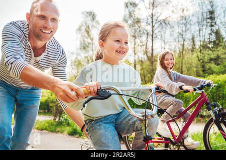 Sorridente padre con due figlie durante una passeggiata all'aperto. Insegna alla ragazza più giovane a fare una bicicletta. Amano stare insieme nel parco estivo della città. Ha Foto Stock