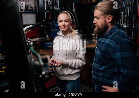 Ritratto di professionista riparatore maschio che comunica con allegro cliente femminile, parlando problema di bicicletta, rilevato durante la diagnostica in riparazione Foto Stock