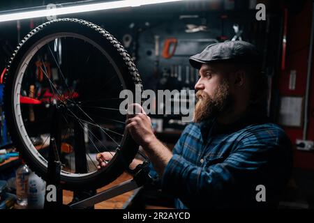 Vista laterale di un riparatore punteggiato in berretto controllando le razze della ruota della bicicletta con chiave a raggi della bicicletta in un negozio di riparazione delle biciclette con interni scuri. Foto Stock