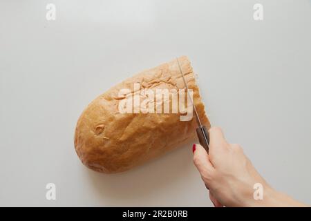 la mano femminile taglia un pezzo di pane con un coltello su un tavolo bianco in cucina a casa Foto Stock