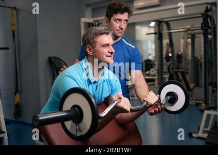 Adulto felice che esegue l'esercitazione di resistenza del bicep con l'addestratore personale Foto Stock