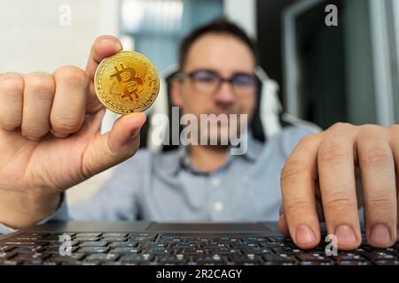 Primo piano scatto di un uomo sorridente che tiene in mano il bitcoin. Business, finanza, concetto di valuta crittografica. buon commerciante con una moneta d'oro del bitcoin al calcolo Foto Stock