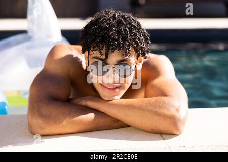 Ritratto dell'uomo felice in forma biraciale in occhiali da sole in piscina appoggiato con le braccia incrociate Foto Stock