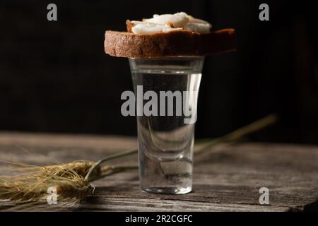 Un bicchiere di vodka accanto a un pezzo di pane nero con pancetta su un tavolo di legno in cucina per il pranzo in ucraino Foto Stock