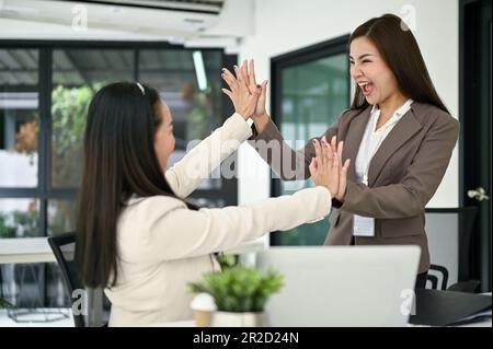 Due giovani donne d'affari asiatiche eccitate e overjoyed stanno urlando con gioia e dando i pomfi alti per celebrare il loro successo di progetto. Concetto di lavoro di squadra Foto Stock