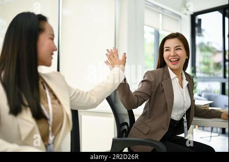 Due giovani donne d'affari asiatiche gioiose ed eccitate, che si danno a vicenda un alto numero di five per rallegrarsi durante il lavoro. Foto Stock