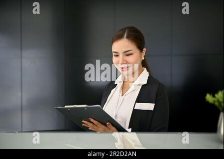 Una bella e fiduciosa receptionist femminile asiatica millennial che legge e controlla un documento alla reception del banco. Foto Stock