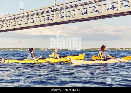 Gruppo di kayak pagaia sotto un ponte al tramonto a casco Bay, Maine Foto Stock
