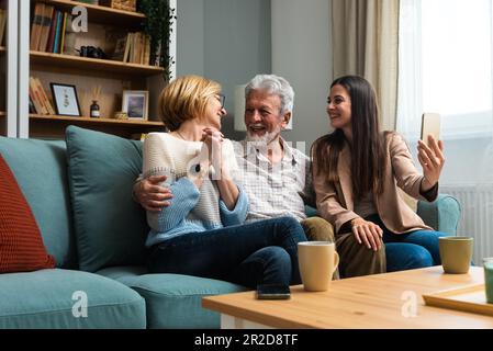 La figlia o la nipote che visitano i suoi genitori, una coppia anziana o i nonni scatta una foto di loro con il suo telefono e prende un selfie come memoria Foto Stock