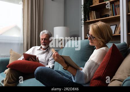 Vita semplice. Felice coppia anziana di mezza età 70s marito e moglie si siedono rilassarsi sul divano in soggiorno leggere libro bere tè insieme, calma anziani Foto Stock