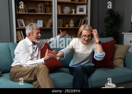 Irritato metà-età o coppia anziana ha duro dibattito a casa, seduta sul divano e discutente, donna infastidita che parla con l'uomo arrabbiato, sposi maturi ha Foto Stock