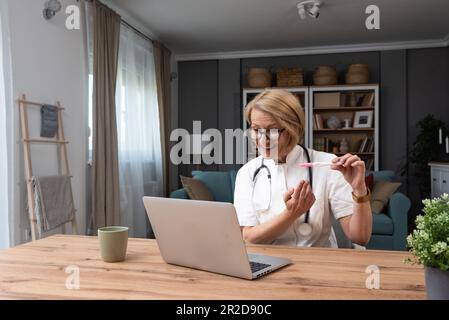 Medico senior esperto che mostra test di gravidanza in videochiamata online sul computer portatile mentre si siede in casa di ufficio da pratica privata in bianco coa Foto Stock