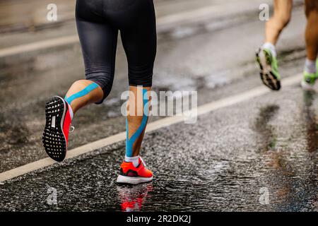 gambe atleta runner femmina correre maratona gara su asfalto bagnato, cinesio nastro blu su shins Foto Stock