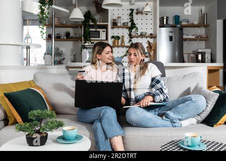 Due giovani donne e i migliori amici che fanno un po 'di shopping online a casa con un computer portatile e una carta di credito da vendita online negozio web mentre sorridono e t Foto Stock